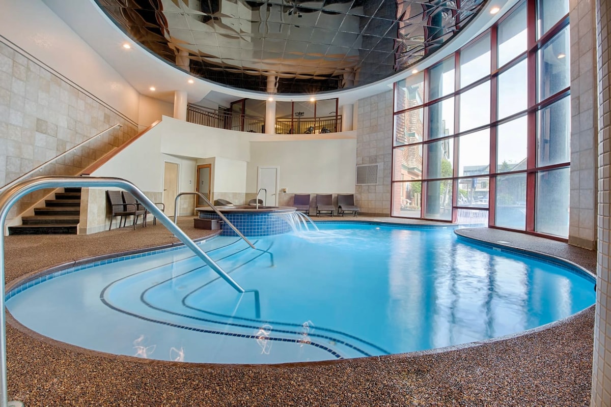An indoor pool area features a curved design with a shallow entry and gentle steps leading into the water. The pool is framed by large windows that allow natural light to fill the space, complemented by modern tilework and seating areas nearby.