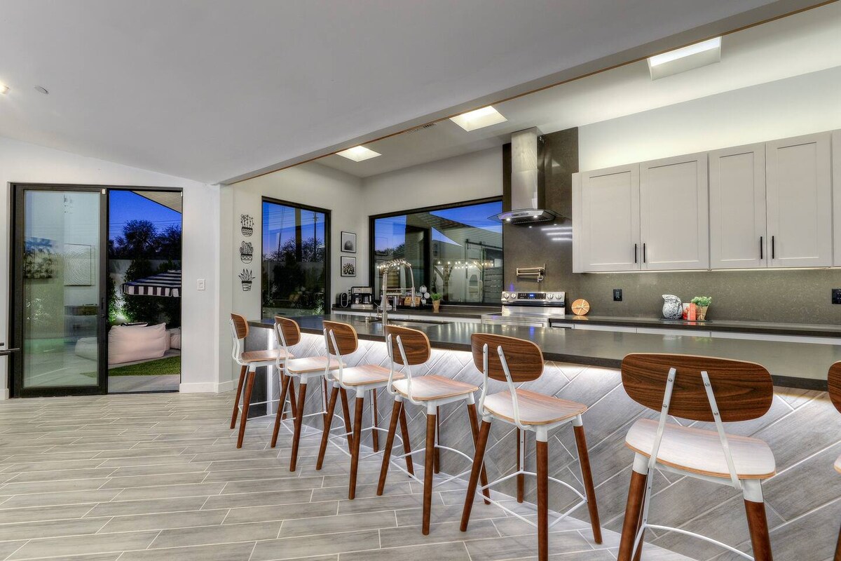 The kitchen features a modern design with a spacious island that seats eight. Large windows invite natural light and provide views of the outdoors. Sleek cabinetry and stainless steel appliances are present, enhancing the functional and stylish atmosphere of the space.