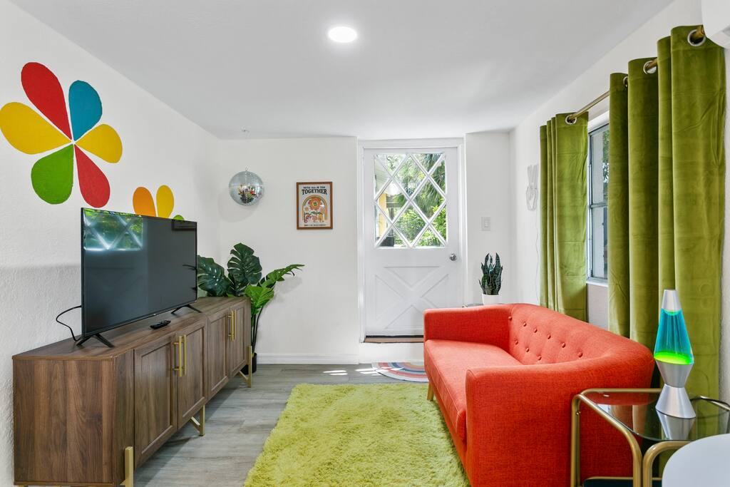 A vibrant living space features a bold orange sofa and a wooden TV console, complemented by colorful wall decor showcasing a flower design. Green curtains frame the windows, while a fluffy green rug adds warmth to the floor. A potted plant and decorative lamp enhance the inviting aesthetic.