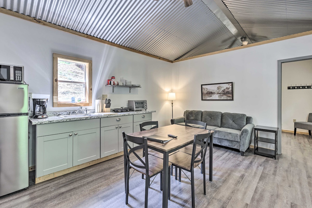 A bright kitchen area showcases modern appliances, including a refrigerator and microwave. A dining table with chairs is positioned nearby, while a comfortable sofa is situated against the wall. Natural light filters through a large window, lending a welcoming ambiance to the space.