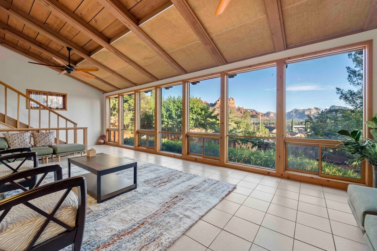 A spacious living area features large windows that provide expansive views of the surrounding red rock landscape. Natural light fills the room, highlighting comfortable seating arrangements and a coffee table set upon a patterned rug. The ceiling showcases wooden beams, enhancing the open atmosphere.