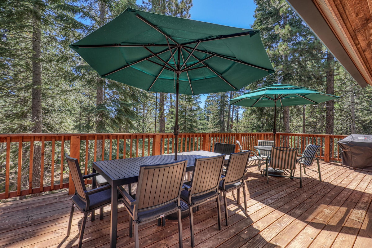 A spacious outdoor deck features ample seating with six dining chairs and a large table, complemented by two shaded umbrellas. Surrounding pine trees provide a natural backdrop, enhancing the outdoor experience for relaxed gatherings.
