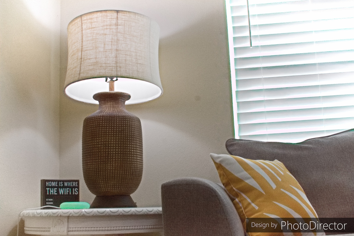 A cozy corner is highlighted by a textured lamp with a soft shade providing warm light. A small decorative sign reading 'Home is Where the WiFi Is' rests on a side table, accompanied by a gray sofa adorned with a patterned yellow pillow. Natural light filters through partially closed blinds.