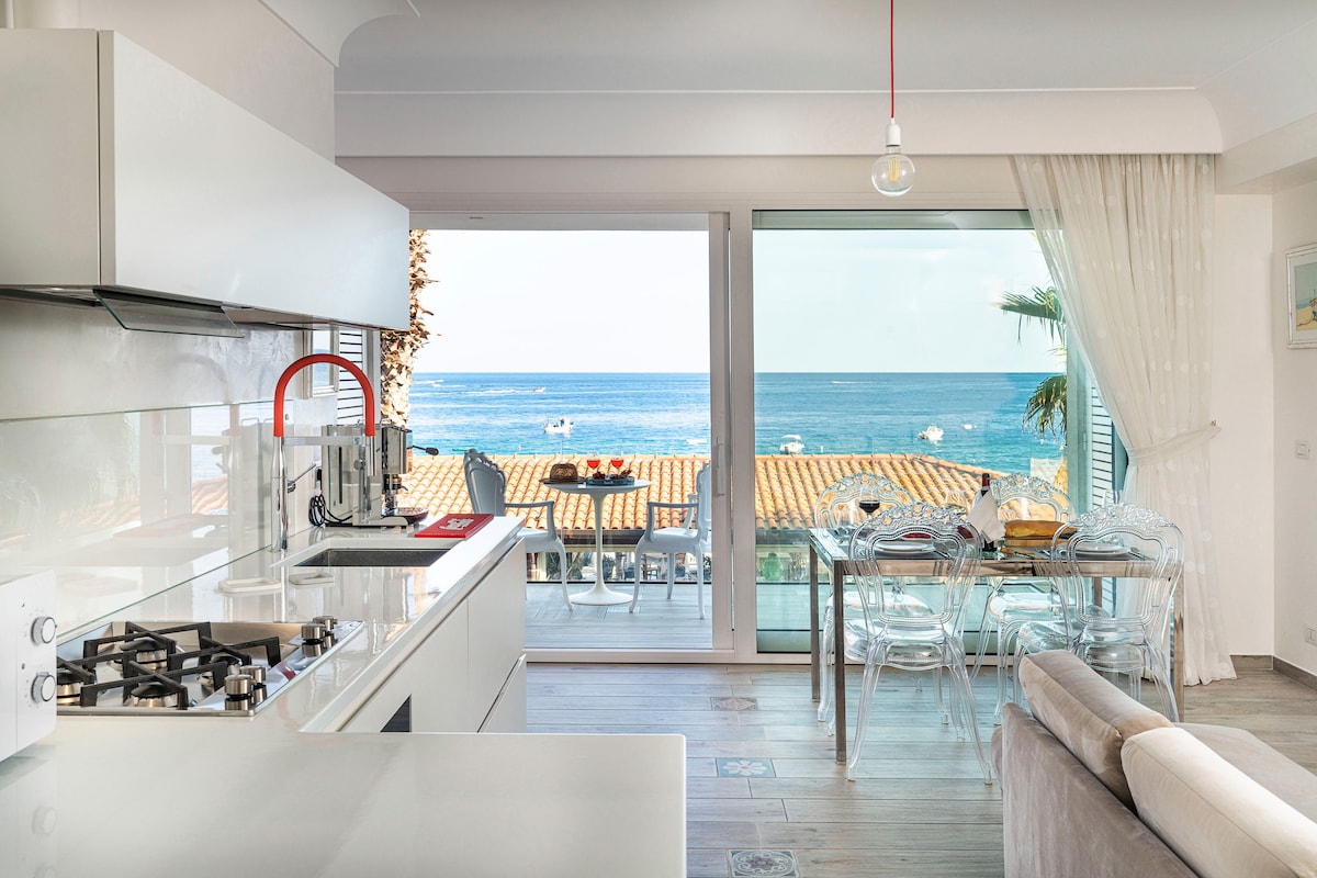 The kitchen and living area feature modern appliances and a sleek design. Large glass doors provide a view of the sea and terrace, where outdoor seating can be observed. Natural light fills the space, accented by light-colored cabinetry and a neutral-toned sofa.