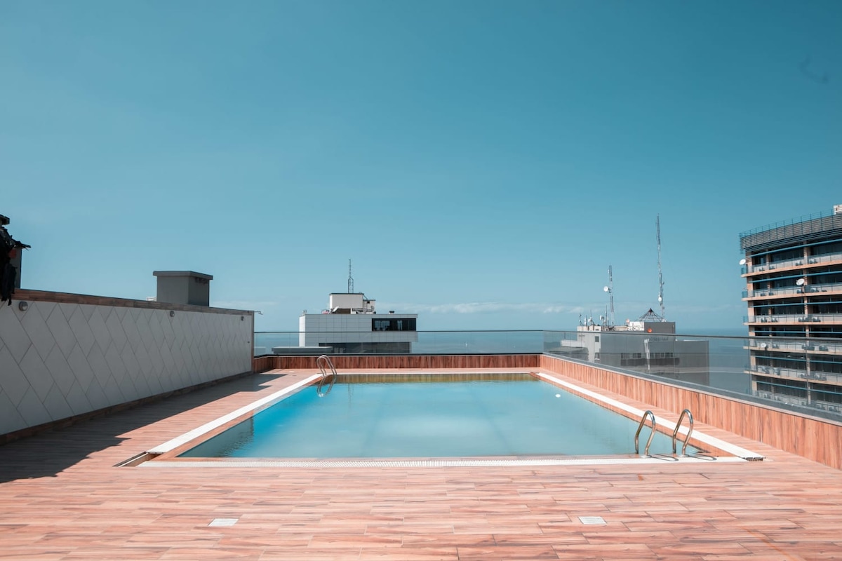A rooftop pool surrounded by a wooden deck offers a refreshing retreat. The clear blue water is complemented by a distant view of the city skyline. A ladder provides access to the pool, and the area is bathed in natural light under a clear sky.