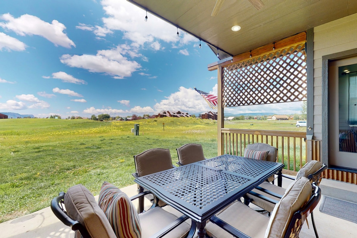 An outdoor patio is furnished with a large table and six cushioned chairs. A pleasant view of open grassy fields and distant mountains is presented, while a decorative lattice feature provides partial shade. The sky is bright with scattered clouds, and an American flag is visible in the background.