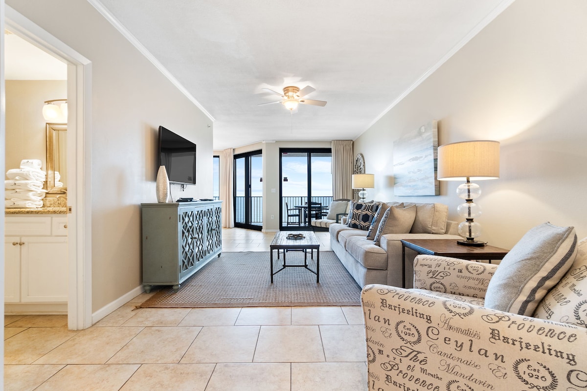 A spacious living area features a comfortable sofa and cushioned chairs, bathed in natural light from floor-to-ceiling sliding glass doors. A large LCD TV is mounted on the wall, while a textured rug and decorative lamp create a relaxed atmosphere. The nearby kitchen and balcony views add to the inviting space.