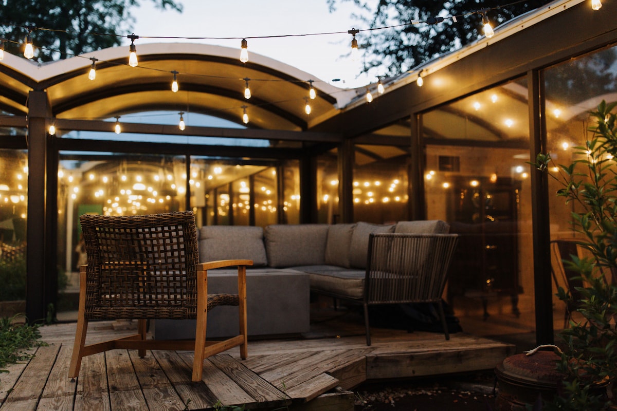 An outdoor seating area is enclosed with glass walls, featuring a sectional sofa and a single chair made of woven material. Decorative string lights cast a warm glow, enhancing the inviting atmosphere for evening gatherings.