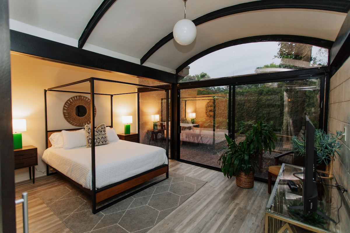 A modern bedroom features a spacious bed with a four-poster frame, complemented by bedside lamps on wooden stands. Floor-to-ceiling windows offer views of the garden outside, while natural light fills the room. A large plant is placed in one corner, adding a touch of greenery.