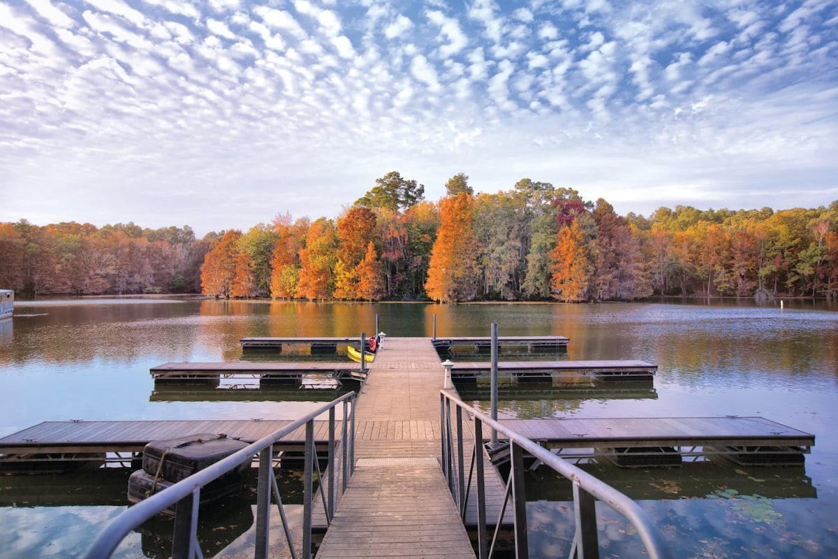 A wooden dock extends over a calm lake, flanked by multiple boat slips. Lush trees in vibrant autumn colors reflect in the water, under a sky filled with scattered clouds. The tranquil scene invites outdoor activities and relaxation.