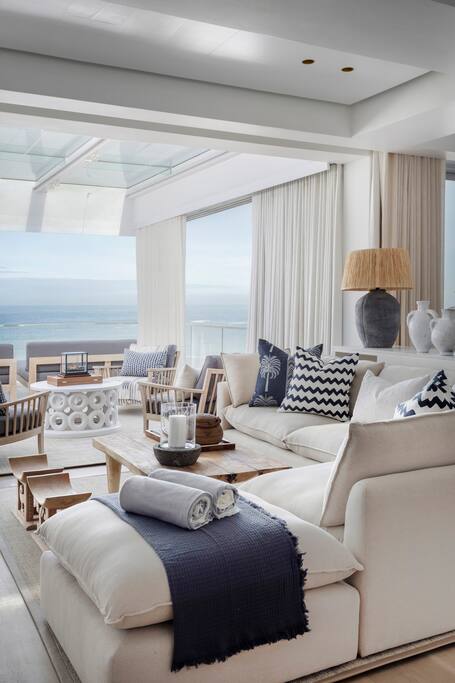 A spacious living area is showcased with a large, comfortable sofa adorned with various cushions and a textured throw. A wooden coffee table sits at the center, surrounded by additional seating. Floor-to-ceiling windows offer expansive views of the sea, bathed in natural light.