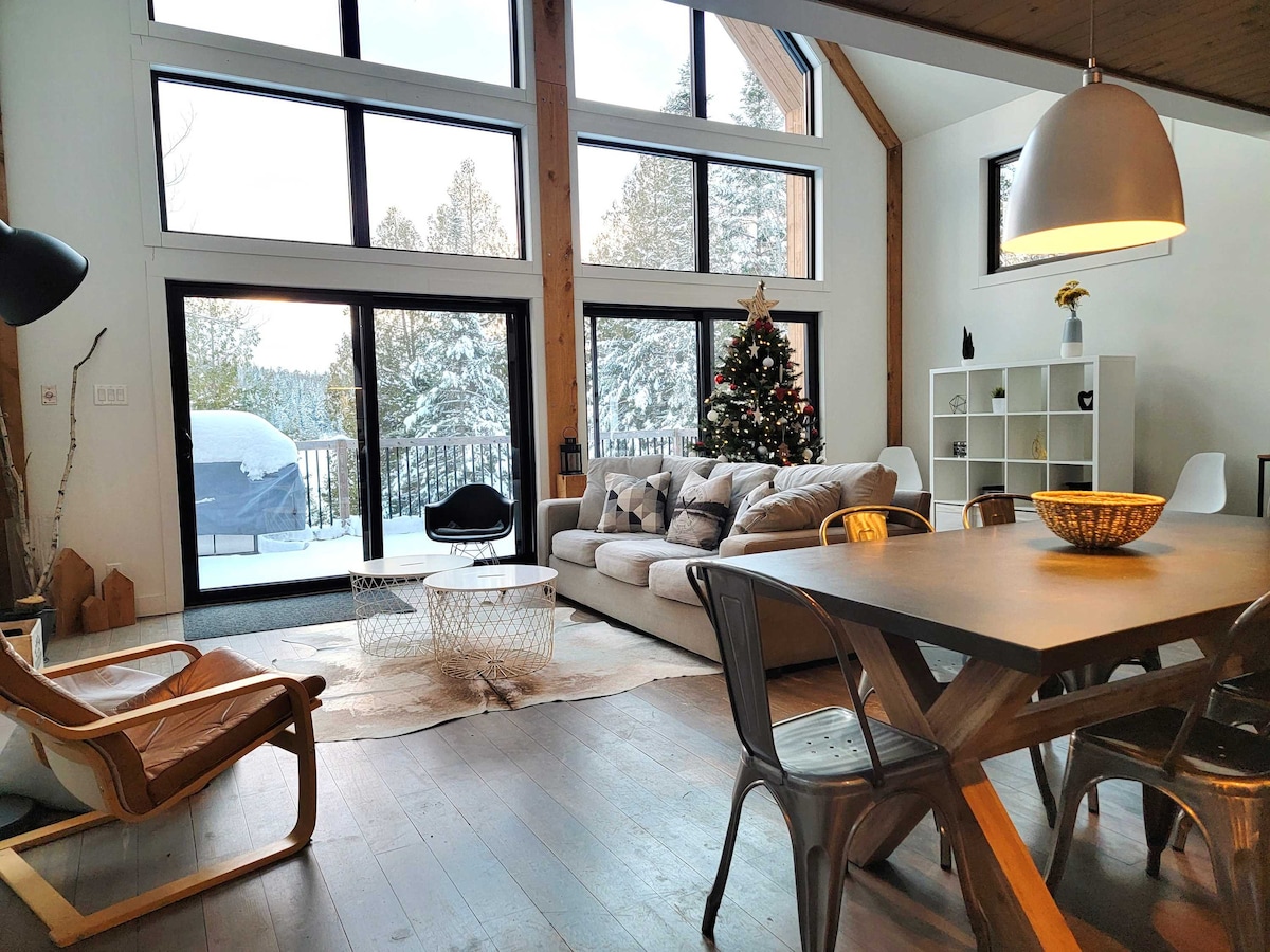 A spacious living area is illuminated by large windows, offering views of snowy trees outside. A plush sofa faces a warm fireplace, accompanied by a dining table and stylish chairs. A Christmas tree adds a festive touch, while a simple decorative bowl sits at the center of the table.