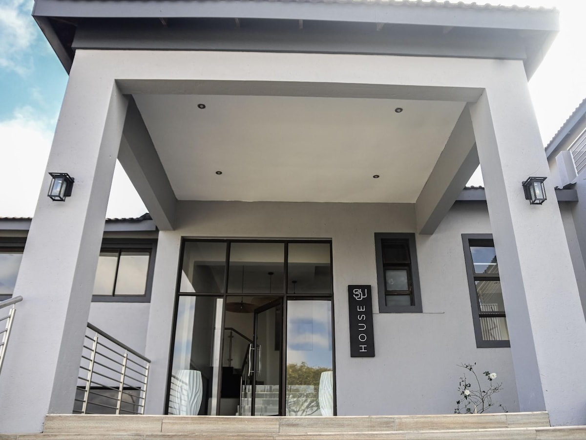 A modern exterior entrance is visible, featuring a spacious covered porch with recessed lighting. Large glass doors lead into the interior of the house, framed by neutral-colored walls. The prominence of the entrance is highlighted by sleek architectural lines and minimalistic design elements.