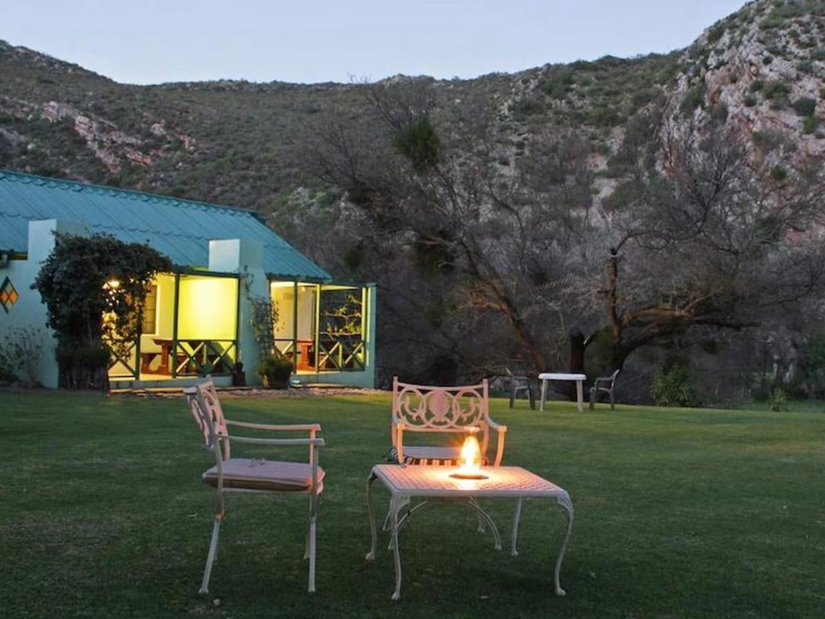 An outdoor area features a small table with a flame in the center, surrounded by two metal chairs. In the background, a green-roofed cottage is visible, with warm light coming from the windows. Lush foliage and mountainous terrain create a serene setting.
