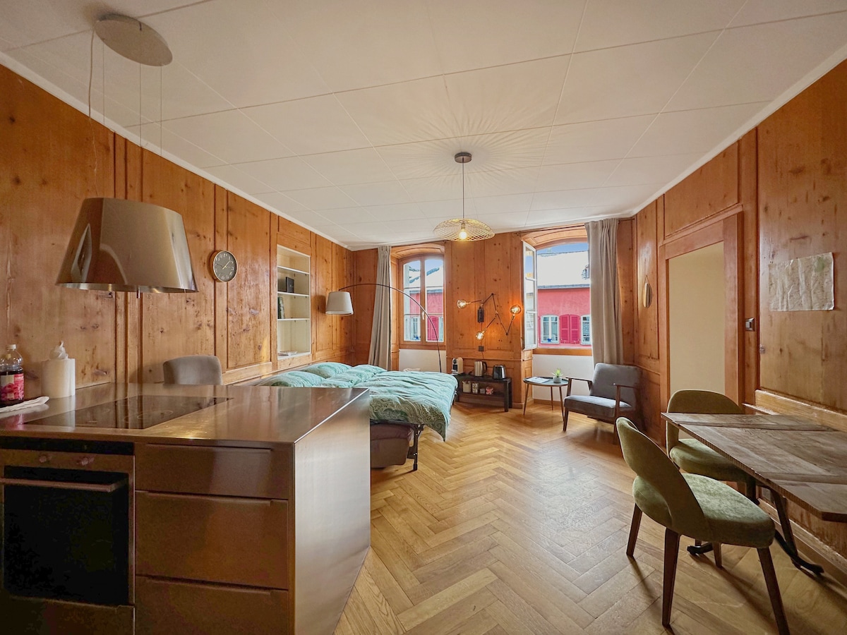 A spacious living area features warm wooden walls and a cozy layout. Large windows allow natural light to fill the room. A comfortable bed with soft bedding is accompanied by a dining table and chairs, along with a kitchenette that provides essential amenities.