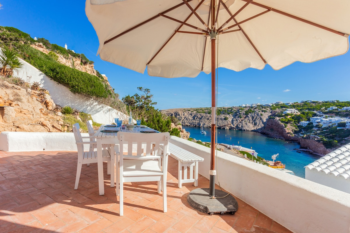 An outdoor terrace is furnished with a dining table and chairs, shaded by a large umbrella. Stunning views of the calm sea and surrounding coastal landscape are visible, accentuating the tranquil setting. The terracotta flooring complements the serene atmosphere.