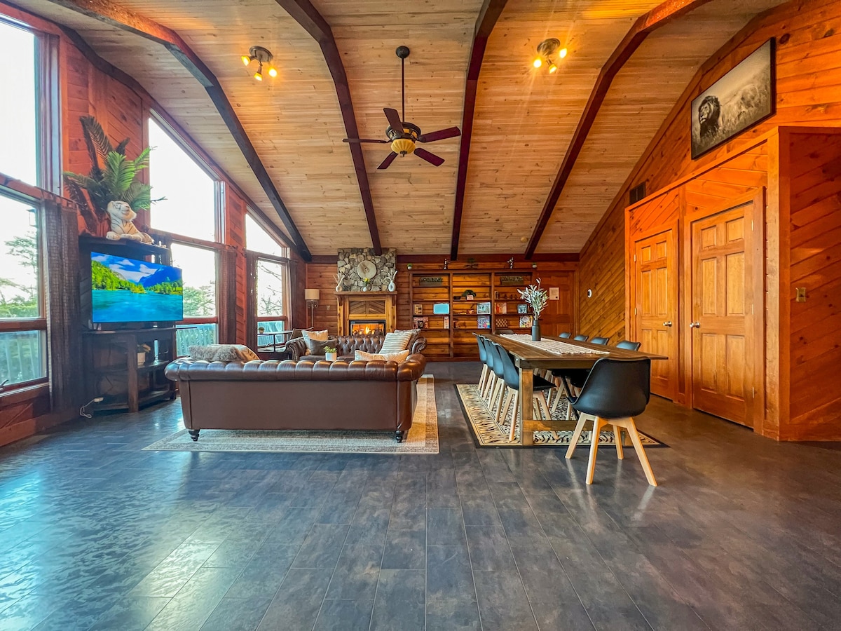 The spacious open-concept living and dining area features high ceilings with wooden beams. A leather Chesterfield sofa and a large dining table set for twelve are visible. Natural light floods in through expansive windows, complemented by a mounted 4K TV and custom wooden cabinetry.