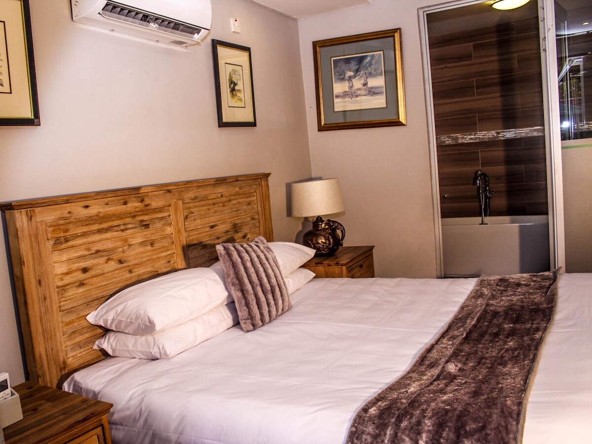 A spacious suite contains a king-size bed with stacked pillows and a decorative throw blanket. Wooden nightstands are positioned on either side, complemented by framed artwork on the walls. A private bathroom with a shower and bath is located in the background.