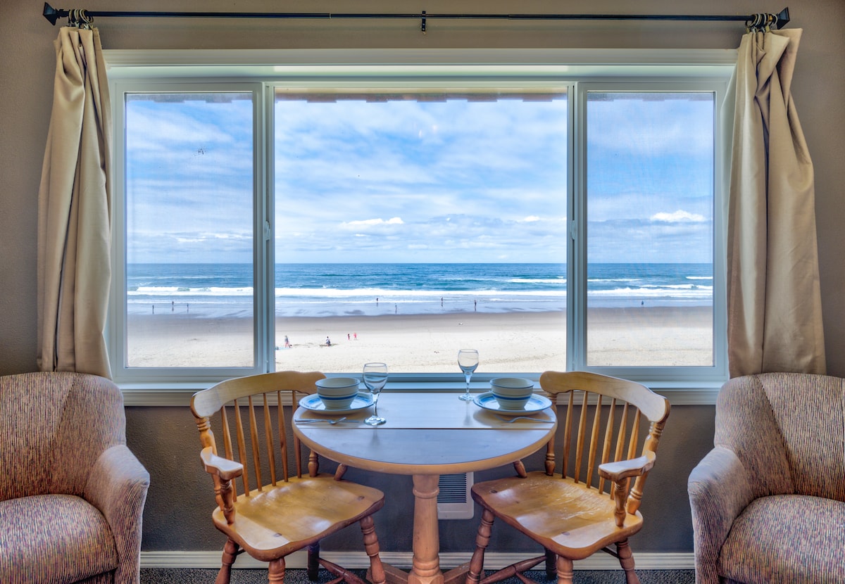 A cozy dining area is positioned beside a large window, offering a view of the ocean and sandy beach. Two wooden chairs surround a round table, set with dishware and glassware. Natural light fills the space, creating a bright and inviting atmosphere.
