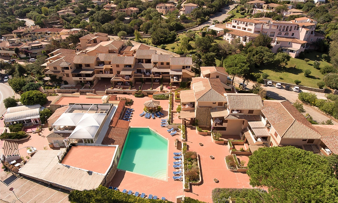 An aerial view of a holiday apartment complex reveals a central swimming pool surrounded by tiled deck areas and lounge chairs. The surrounding buildings feature Mediterranean-style architecture with terracotta roofs and landscaped gardens, while pathways connect various units within the complex.
