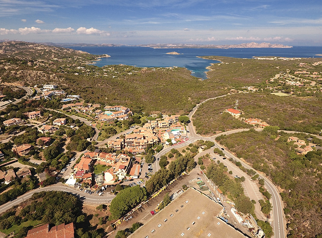 An aerial view of the Porto Cervo Marina area, featuring a cluster of buildings surrounded by lush greenery. The coastline and turquoise waters can be seen in the distance, highlighting the scenic landscape that blends nature with the resort's accommodations.