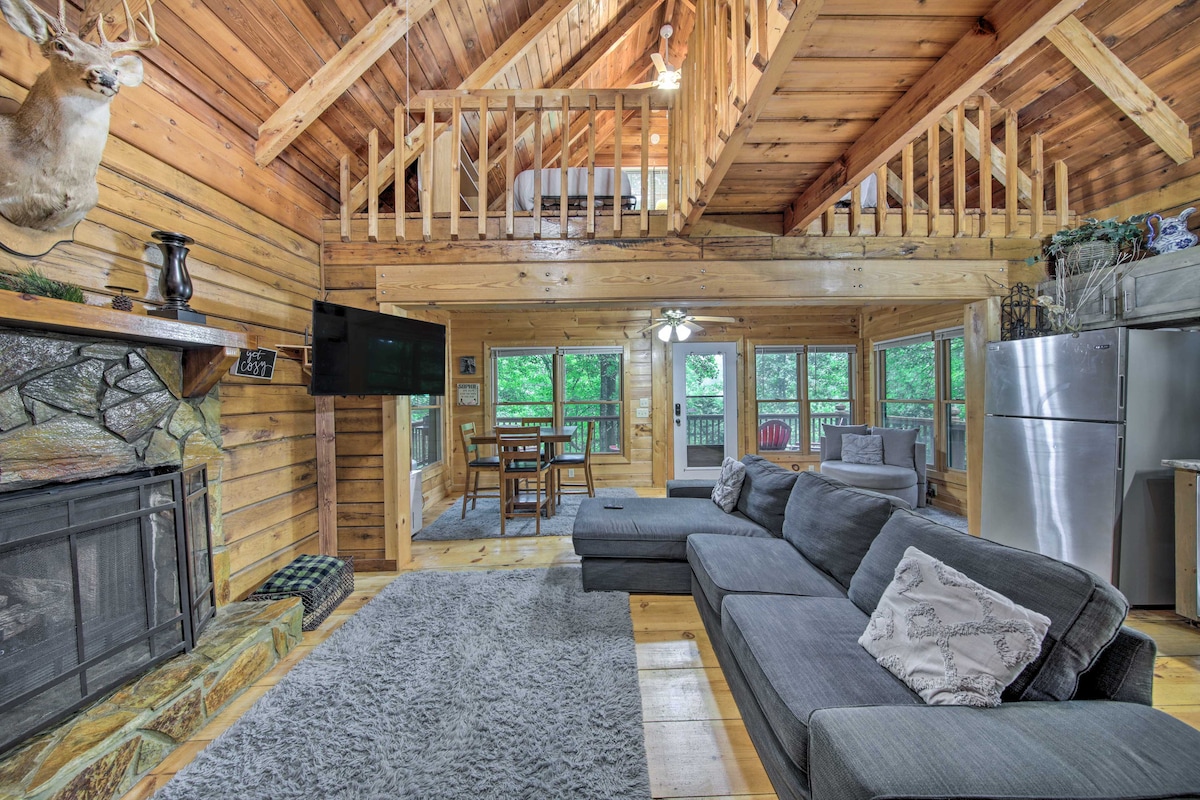 A spacious living area is highlighted by a large sectional sofa facing a stone fireplace. Large windows provide natural light and views of the surrounding trees. A dining table is visible on the loft level, and modern kitchen appliances complement the rustic wooden interior.