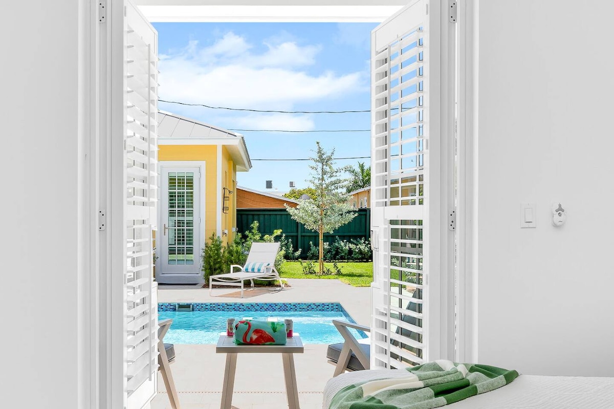 Double doors open to reveal a private pool area with a sparkling blue pool surrounded by lush greenery. The view includes lounging chairs and a bright yellow pool house in the background, inviting relaxation in a sunny atmosphere.