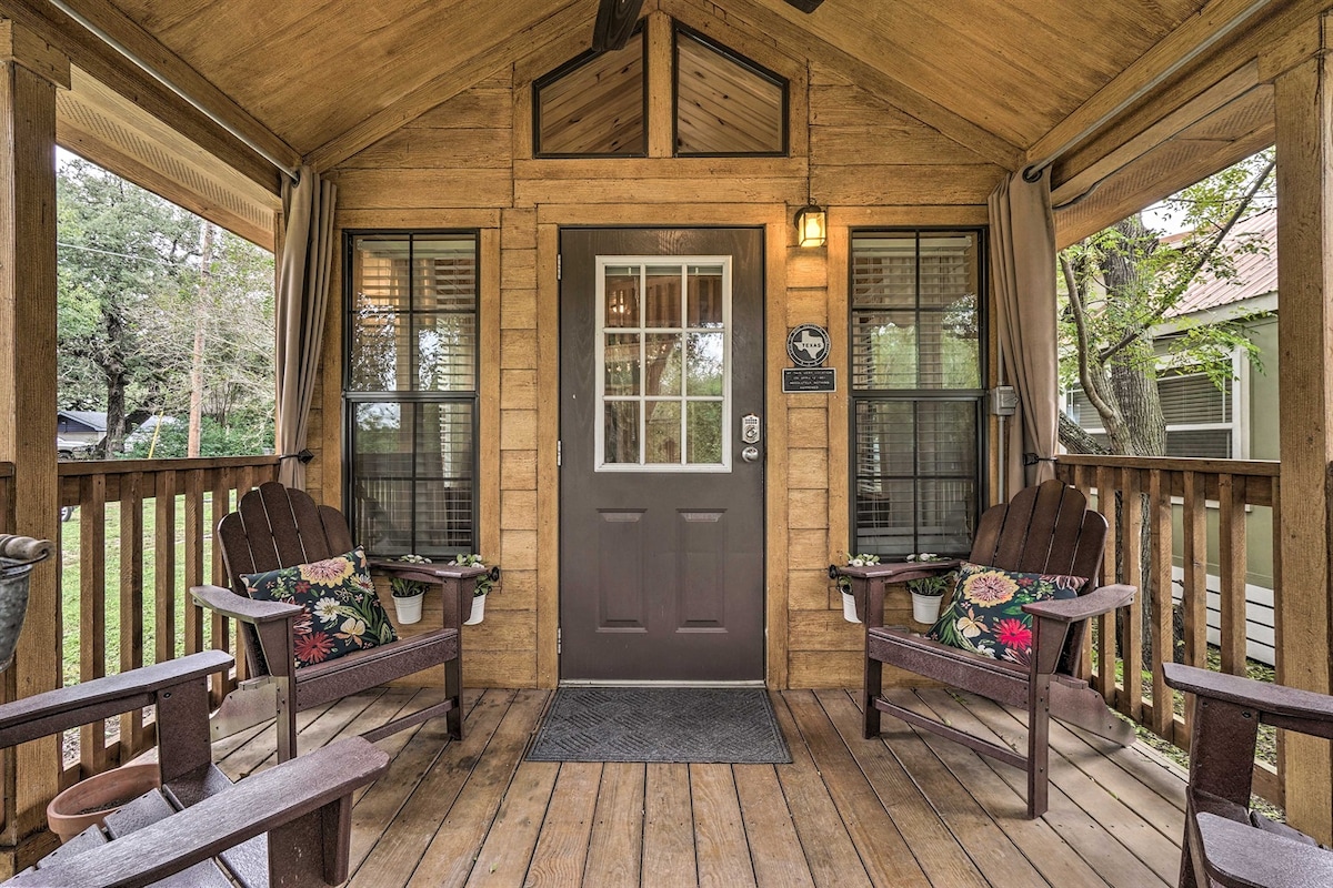 An inviting porch features wooden flooring and is furnished with two adirondack chairs adorned with floral cushions. A front door with a window is positioned centrally, flanked by large windows that offer views of the surrounding greenery.