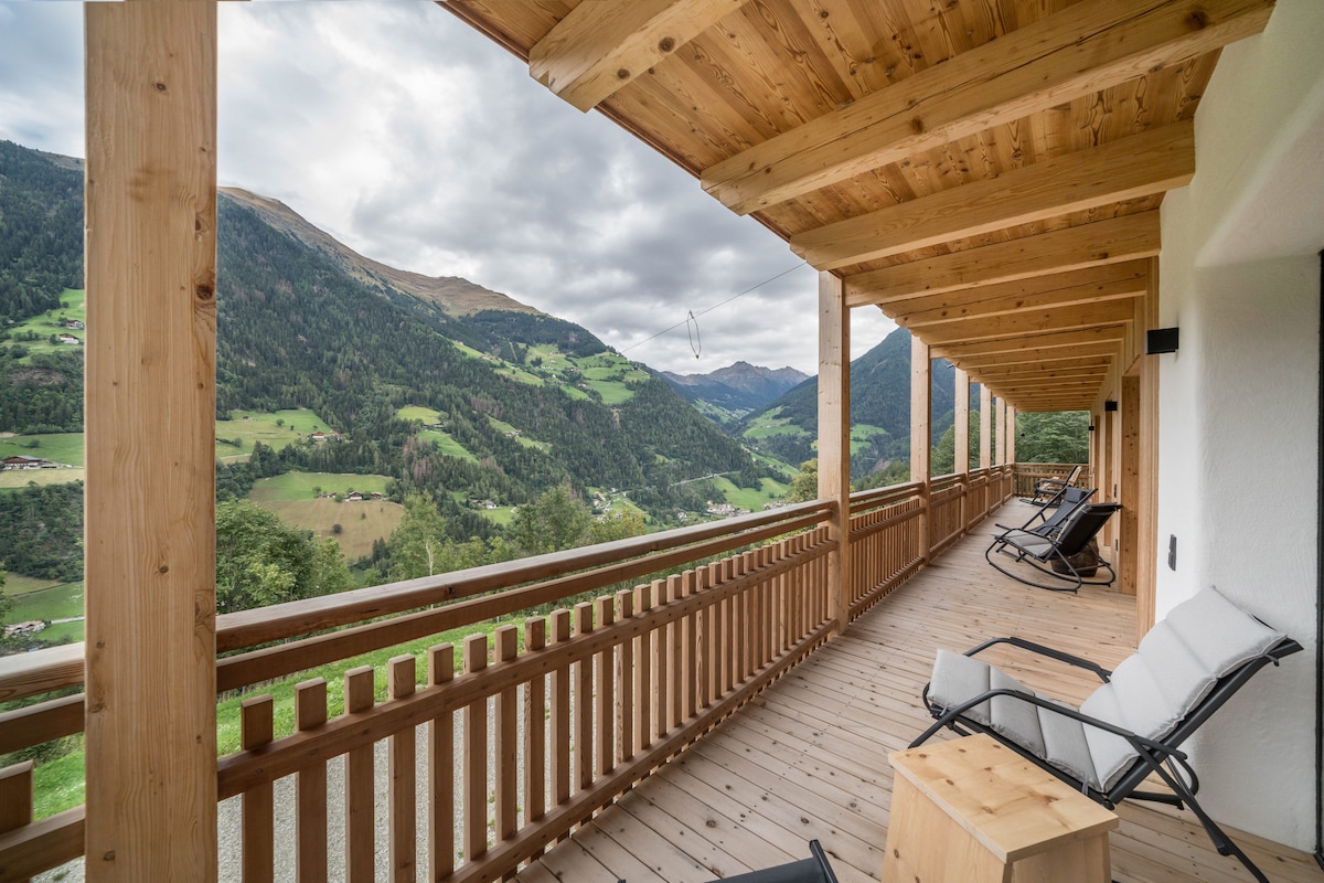 A spacious wooden terrace is furnished with comfortable lounge chairs, offering panoramic views of the surrounding mountains and valleys. The natural wood structure enhances the connection to the outdoors, while the open space allows for relaxation and enjoyment of the scenery.