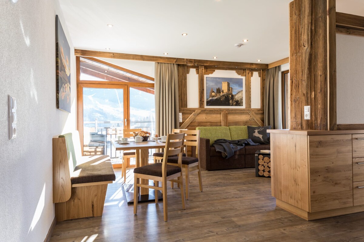 A spacious living area features wooden accents and a cozy seating arrangement, including a sofa and a dining table. Large glass doors lead to a balcony, allowing natural light to fill the room. Scenic mountain views are visible through the curtains.