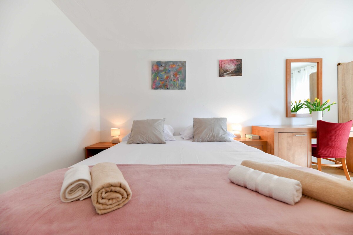 A serene bedroom is presented, featuring a spacious bed with soft linens. Two lamps provide gentle lighting on either side. A desk with a mirror is positioned nearby, complemented by a red chair. Fresh flowers add a touch of color to the space.