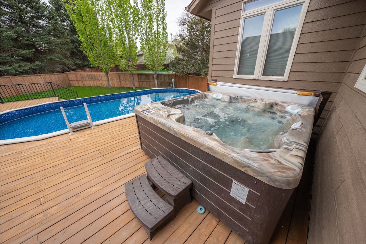 A six-person hot tub is positioned on a wooden deck beside a heated pool. The pool's blue water is visible, surrounded by a wooden fence, providing privacy. Lush green trees can be seen in the background, creating a serene outdoor setting.