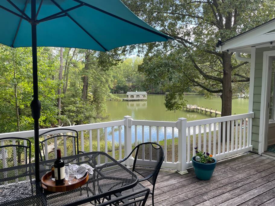 A deck view reveals a serene cove surrounded by lush greenery. The outdoor dining set, complete with an umbrella, overlooks calm waters and a distant dock. The scene captures a tranquil atmosphere, inviting guests to enjoy meals or relaxation outdoors.
