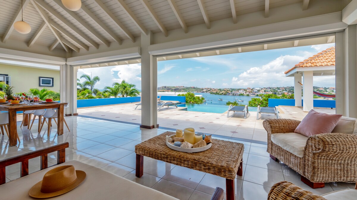 The spacious living area features large glass doors that open to a scenic view of Spanish Water and the Caribbean Sea. A comfortable seating arrangement is positioned in front of a rustic coffee table, with a dining table set for meals. Sun loungers are visible on the terrace.