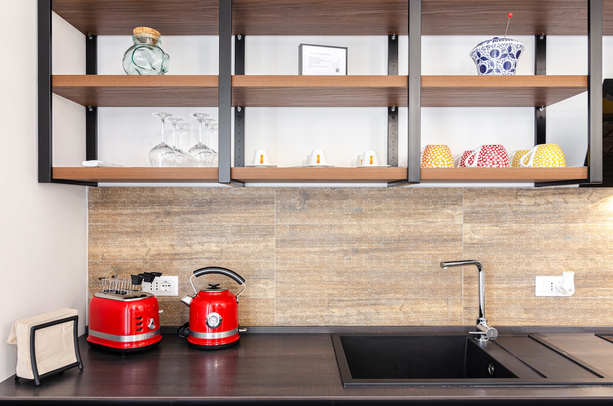 The kitchen features a black countertop with a sleek sink and two red appliances—one toaster and one kettle. Above the countertop, open shelving holds glassware and colorful mugs, adding vibrancy to the space. The warm-toned wall creates an inviting atmosphere.