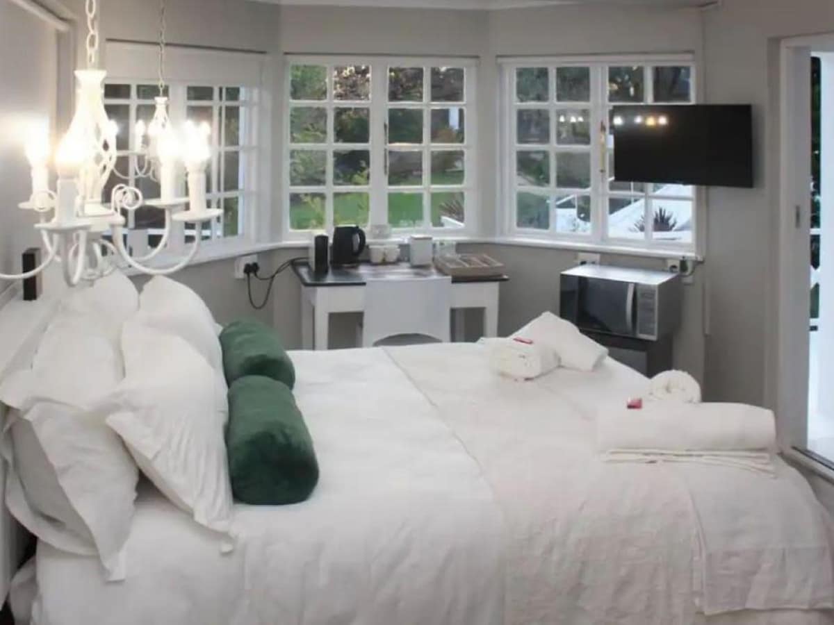A well-lit room features a spacious bed adorned with white linens and green accent pillows. Large windows provide ample natural light, and a small kitchenette with a kettle and microwave is visible. A flat-screen TV is mounted on the wall, enhancing the room's amenities.