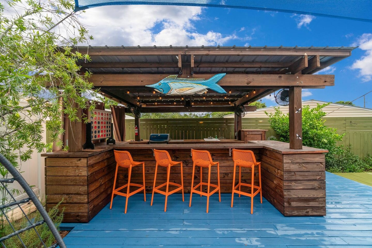 An outdoor bar area features a wooden structure with a large, fish-themed decoration above the counter. Orange bar stools are positioned around the counter, providing vibrant seating options. The space is surrounded by greenery, with blue decking that enhances the inviting setting.