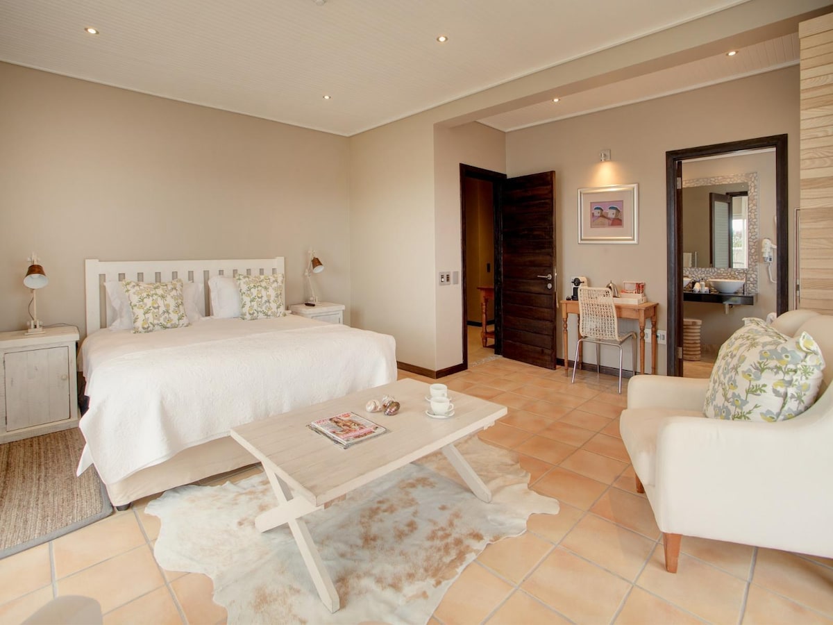 A spacious room features a king-sized bed adorned with decorative pillows. A cozy seating area includes a light-colored chair and a low table. Warm lighting enhances the neutral-toned walls, and an entryway leads to the en-suite bathroom visible in the background.