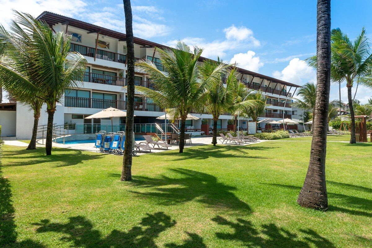 A spacious condo complex is set amidst lush green lawns, with palm trees scattered throughout. Lounge chairs are positioned around the pool area, accompanied by umbrellas for shade. The balconies of the upper floors overlook the pool, contributing to the serene beachfront atmosphere.