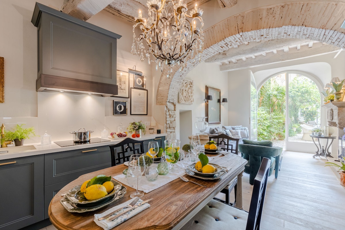 A modern kitchen and dining area with an elegant chandelier, highlighting a wooden dining table set with tableware and fresh lemons. Natural light floods through an arched window, offering views of the garden. Textured walls and exposed wooden beams create a cozy ambiance.