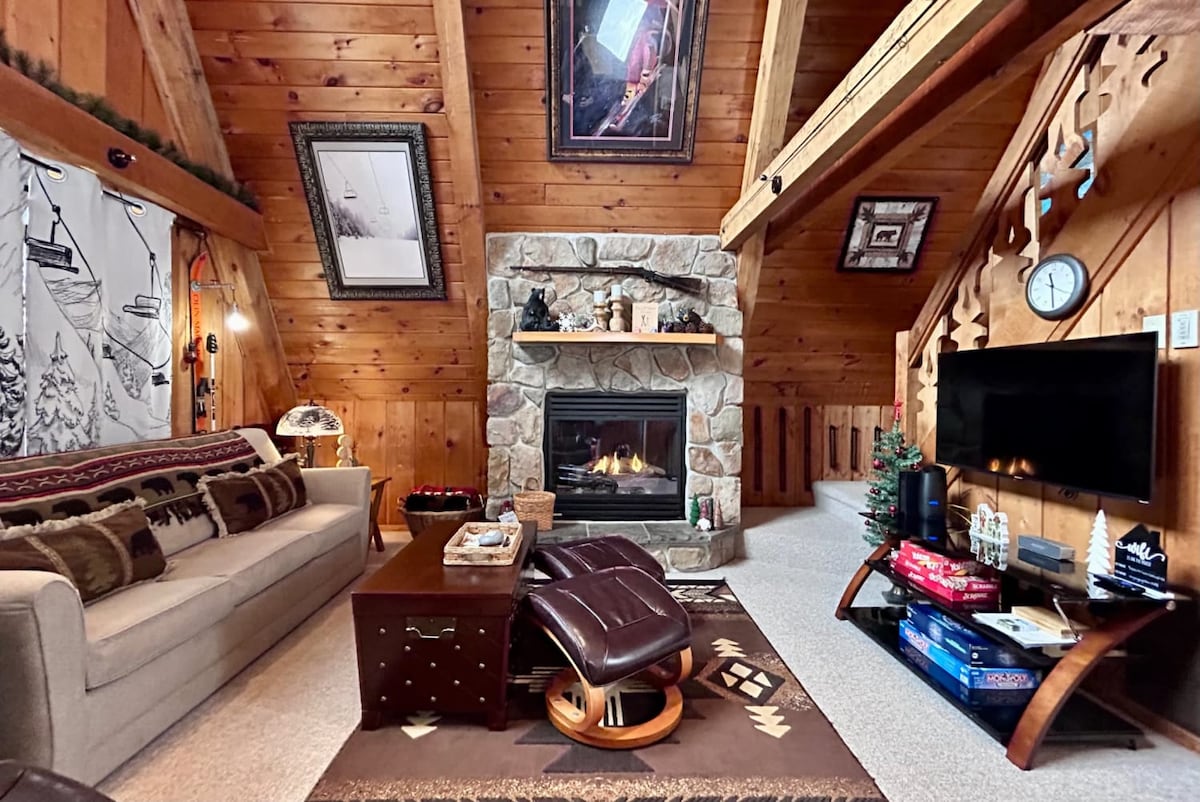 A warm living area features rich wood paneling and a stone fireplace at its center. A light-colored sofa and a dark coffee table create a cozy seating arrangement. Decor includes framed art and a clock on the wall, enhancing the inviting space.