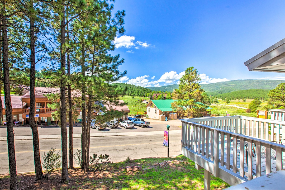 A view from the spacious balcony captures picturesque mountain landscapes and nearby structures. Tall pine trees frame the scene, while cars are parked along the road below. Expansive blue skies with scattered clouds create a vibrant backdrop to the tranquil setting.