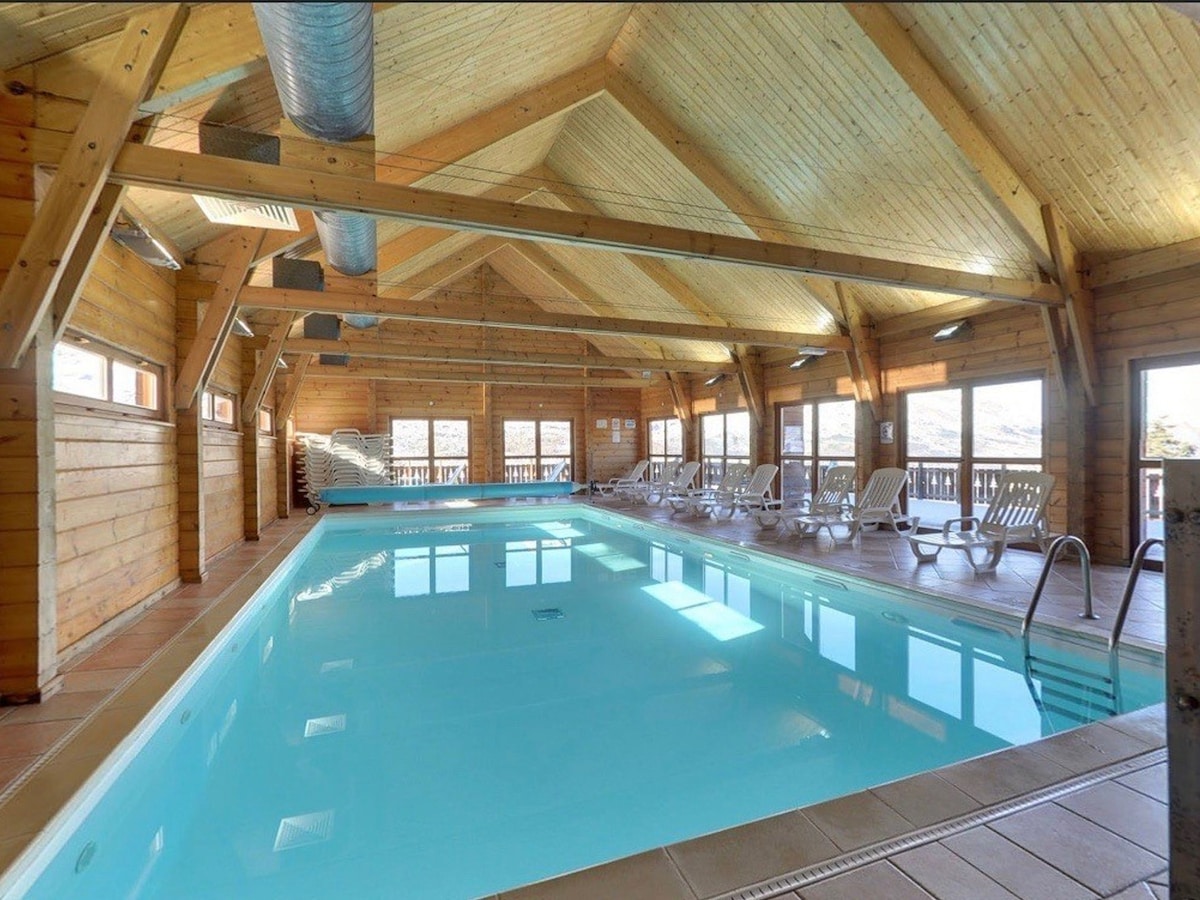 An indoor swimming pool is visible, featuring a spacious layout with clear blue water. Sunlight filters through large windows, illuminating the wooden beams and warm interior. Several lounge chairs line the poolside, providing a comfortable area for relaxation.
