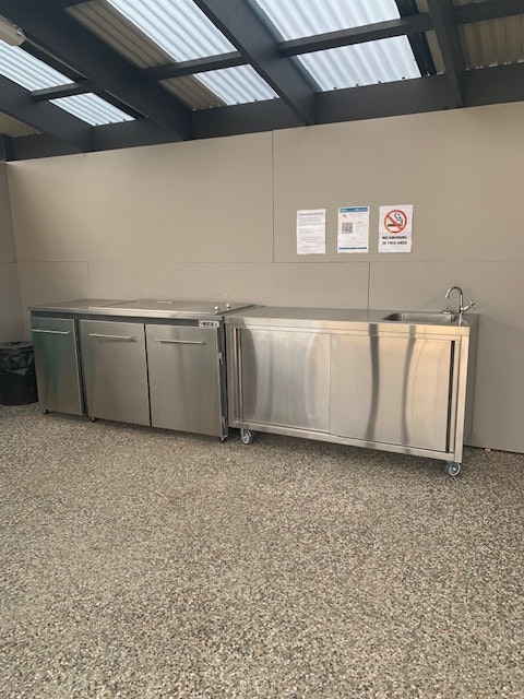 A modern kitchen area features stainless steel cabinets and countertops with a sink. The space is designed with a hard flooring surface, and wall-mounted documents are visible, including a notice sign. The overall design supports a clean and functional cooking environment.