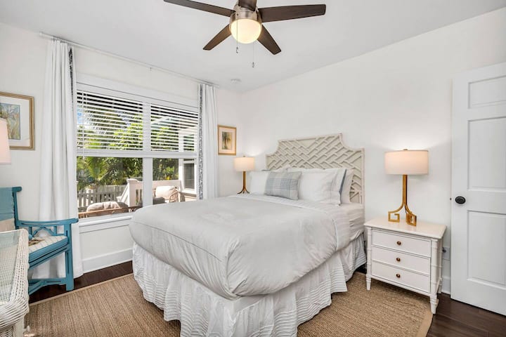 Este hermoso dormitorio cuenta con un tema más costero con su cómoda cama tamaño queen y su cabecera y tocadores blancos de ratán. Disfruta de las elegantes mesas auxiliares con lámparas laterales art déco, una televisión inteligente en streaming y un sillón de color verde azulado brillante.