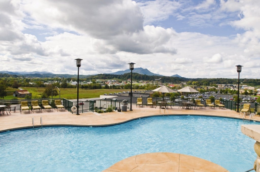 An outdoor pool is visible, surrounded by lounge chairs and umbrellas. Scenic views of gentle green slopes and distant mountains are captured under a partly cloudy sky. The area features decorative lighting fixtures along the edge, enhancing the inviting atmosphere.