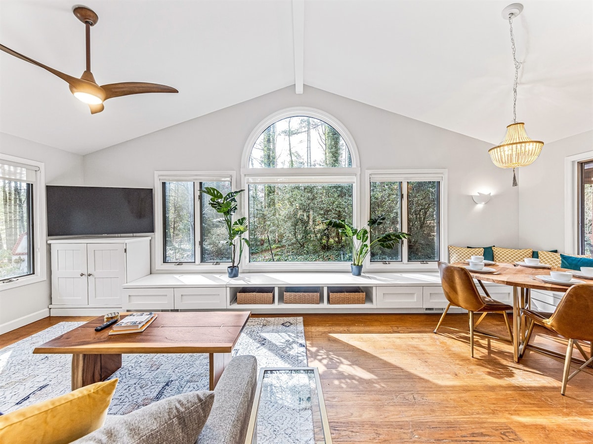 A spacious living area is illuminated by natural light filtering through large windows. A cozy seating arrangement features a gray sofa and a wooden coffee table. The adjacent dining area includes a table set for six, complemented by warm, earthy tones and greenery from potted plants nearby.