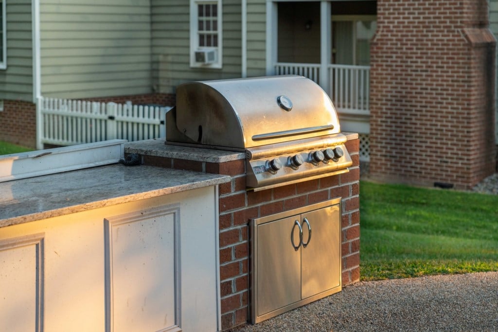 An outdoor grill is situated on a stone countertop, featuring multiple burners and a spacious cooking area. Natural light reflects off the metal surface, highlighting its polished finish. The background includes a lawn area and a glimpse of a nearby building.