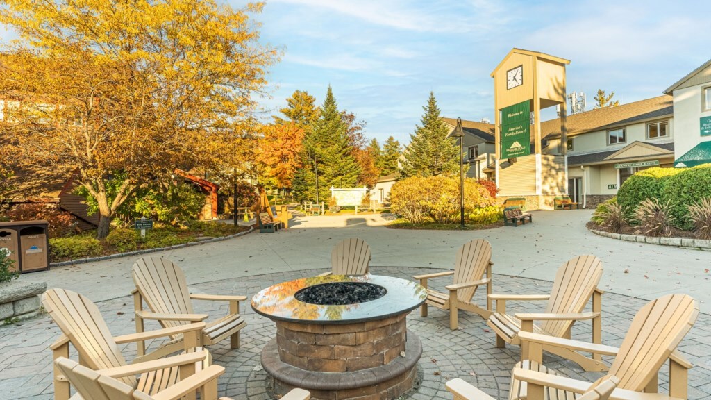 An outdoor gathering space features a circular stone fire pit surrounded by six wooden Adirondack chairs. The area is landscaped with colorful autumn foliage, and a clock tower stands in the background, indicating a welcoming community atmosphere.