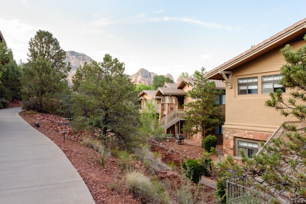 The pathway leads through landscaped grounds featuring native vegetation, with terracotta-hued buildings on either side. The natural surroundings include rocky formations in the background, providing a serene setting for the resort's architecture.